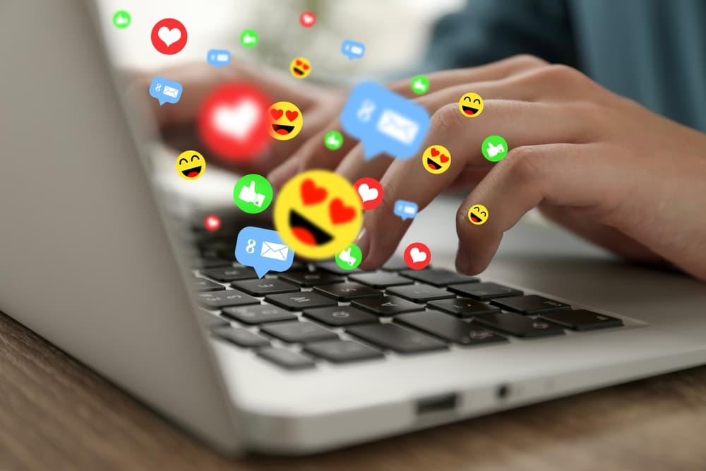 Woman using laptop at table, closeup. Social media notifications (likes, messages and emojis) flying out of computer