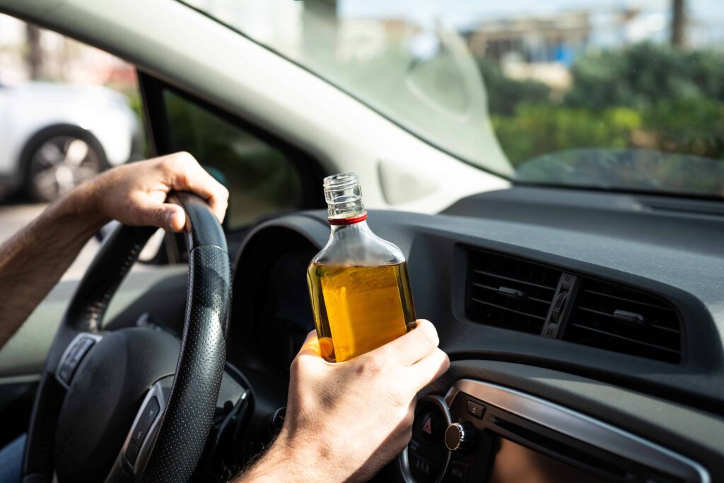 Driver holding an open alcohol bottle while driving, illustrating drunk driving danger.