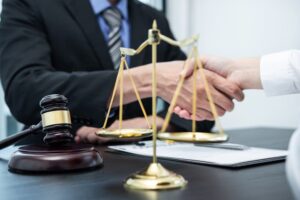 Attorney and client shaking hands across a desk with legal documents, gavel, and justice scales symbolizing a car accident settlement agreement.